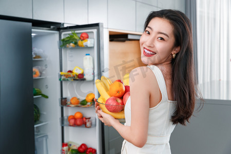 冰箱内的水果摄影照片_拿着水果的青年女人站在冰箱前