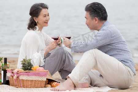 海滩游玩摄影照片_幸福的老年夫妇坐在海滩上野餐饮酒