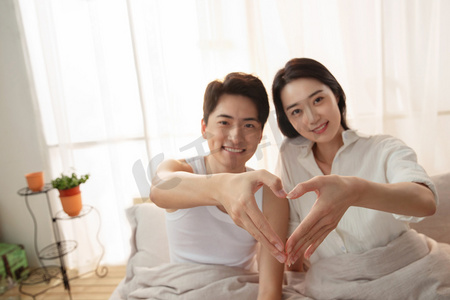 年轻亚洲家庭摄影照片_快乐的年轻情侣在床上做心形手势