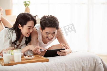 年轻情侣在床上一边吃早餐一边看平板电脑