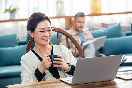 捧着杯子摄影照片_中老年女士在家捧着杯子看笔记本电脑