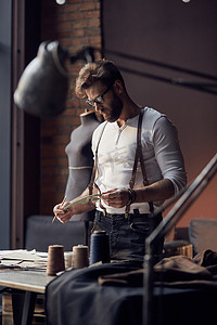 年轻的严重裁缝，留着胡子，穿着白衬衣与寻找附近的木桌和古董家具与模特的惊人工作室中的线程在背景上的绘图上的皮制背带眼镜