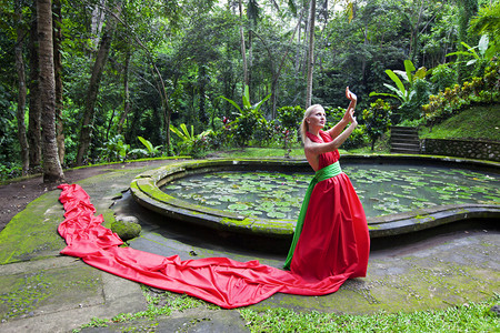 美丽的女孩在一个红色的长礼服站在 waterpool 附近的魔法森林和抵抗风