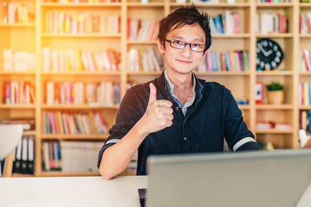 亚洲年轻成年男子与笔记本电脑，ok 的手势，家庭办公室或图书馆场景，竖起大拇指用时钟书架模糊背景与副本空间、 成功或技术的概念、 温暖的光效果