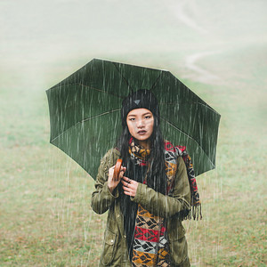 戏剧的女孩拿着雨伞的亚洲外观。雨淋。一个女孩站在早期的天气中的字段的背景。图像的整体计划 / 中期计划。那个女孩看着镜头.