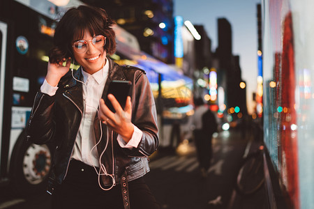 行动电话摄影照片_开朗漂亮的年轻女子在凉爽的眼镜和时髦的穿着步行在大都会街与夜灯享受歌曲从播放列表在耳机和阅读 sms 与好消息智能手机