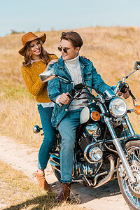 年轻幸福夫妇坐在复古摩托车在乡村草地上