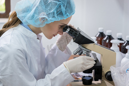 在化妆品厂实验室工作中, 在显微镜下分析样品时佩戴无菌安全设备的女化学家的侧面观察