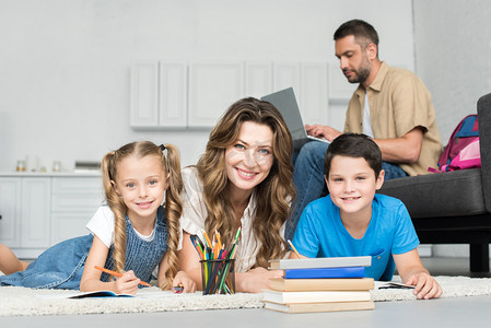 微笑的母亲和孩子们看着相机, 同时做家庭作业和人在沙发上使用笔记本电脑在家