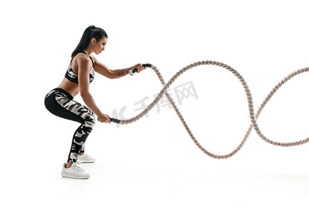 动态白色背景摄影照片_强壮的肌肉发达的女人用战斗绳索工作。在白色背景下的时尚运动服中有吸引力的女人的照片。力量和动力。侧面视图.
