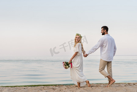 两人牵手摄影照片_快乐的新娘和新郎牵手走在沙滩上的婚礼花束