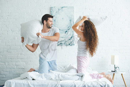 快乐的年轻夫妇的侧面观看睡衣与枕头在床上打架  