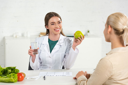 微笑营养师在白色外套拿着苹果和一杯水和病人在餐桌上