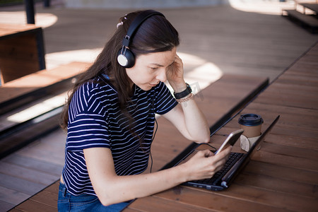一个带着智能手机的女学生，坐在笔记本电脑前，用耳机仔细听，学习外语.