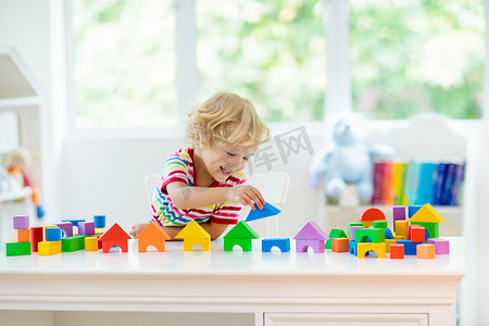 幼儿园家摄影照片_孩子玩彩色玩具块。小男孩用方块玩具建造塔楼。幼儿教育和创意玩具和游戏。婴儿在白色的卧室里用彩虹砖。在家的儿童.