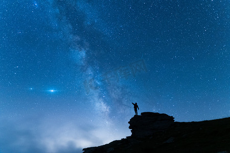 一个站着的年轻人在山上高举双臂的轮廓。带着蓝夜星空旅行的背景.