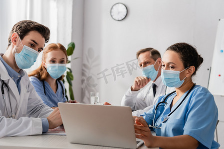 戴着医疗面具的多文化医院工作人员坐在医院的工作场所，在模糊的背景下看着笔记本电脑
