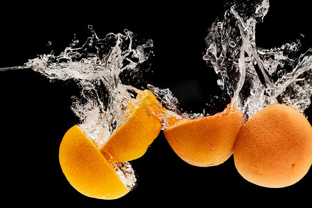 橙子水摄影照片_成熟的葡萄柚和橙色的两半落水，溅到黑色