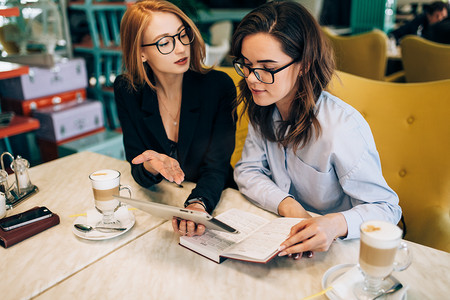 褐红摄影照片_两个女孩在一家咖啡厅的商务会议上。喝咖啡。讨论未来的公司及其项目。签订合同。红头发的女孩和褐发女郎.