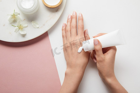 裁剪视图的妇女应用奶油，手牵手靠近盘子与化妆品和鲜花在白色粉红色表面