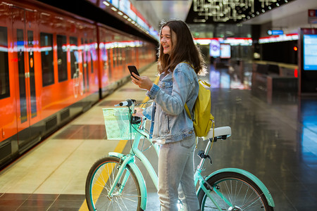 背着背包和自行车的少女站在地铁站，手里拿着智能手机，滚动着发短信，微笑着笑。未来明亮的地铁站。芬兰， 埃斯波