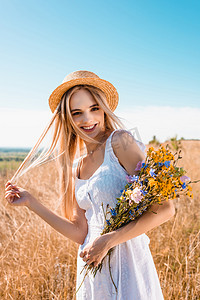年轻时髦的女人，戴着草帽，身穿白色衣服，手持野花，一边看相机一边摸着头发