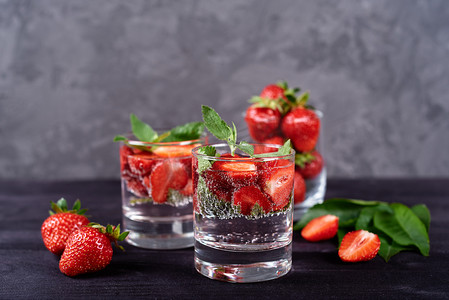 将草莓和薄荷糖倒入水中，用闪闪发光的玻璃杯在木黑色桌子的背景上，复制空间。寒冷的夏季饮料。矿泉水