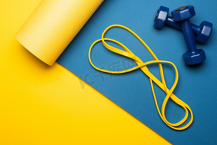 蓝色哑铃摄影照片_带黄色背景哑铃和电阻带的蓝色健身垫顶部视图