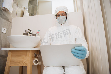 在洗脸盆附近的浴室里，穿着危险垫套装、医疗面罩、乳胶手套和护目镜的男人正在使用笔记本电脑 