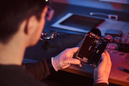 智能照明摄影照片_修理工仔细检查智能手机内部元素的完整性在一个现代化的修理车间。红色和蓝色灯光照明