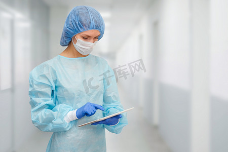 医疗帽子摄影照片_身着防护服的女医生：医疗面罩、帽子和长袍。这名妇女使用数字平板电脑，并在屏幕上显示疾病统计数据.