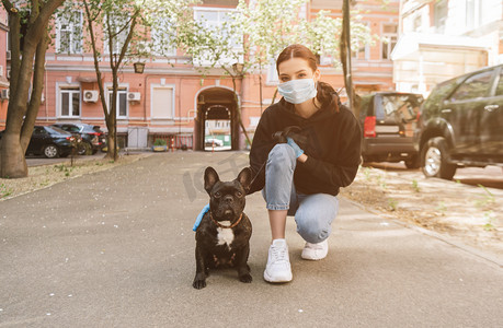 狗绳摄影照片_戴着医疗面罩和乳胶手套在街上摸可爱的法国斗牛犬的女人 