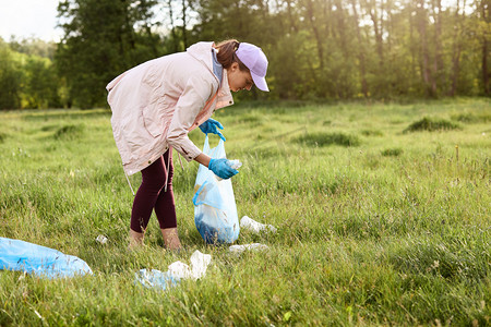 捡垃圾摄影照片_落日时，妇女站在田野中间，一边是散落的布，一边是提着垃圾袋，一边是捡垃圾。环境污染概念，收集垃圾.
