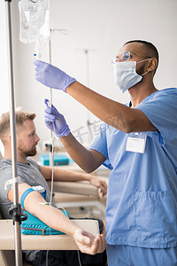 Young mixed-race doctor in blue uniform, protective mask and rubber gloves preparing dropper for one of patients sitting near by