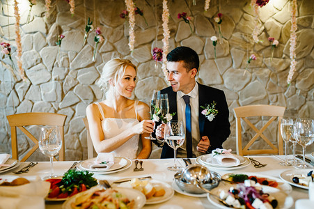 新婚夫妇举着香槟酒在屋里举杯敬酒。用鲜花和绿色装饰的婚礼仪式。餐厅宴会厅餐桌旁的新郎新娘.