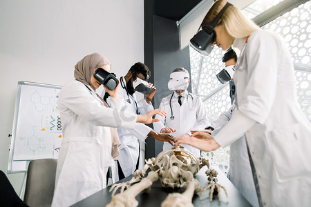 model摄影照片_Team of multiethnic professional doctors, surgeons, orthopedists, students wearing VR goggles working with human skeleton model in modern classroom. Virtual anatomy, medicine concept