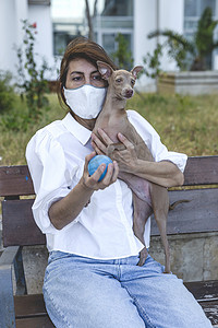 抵御严寒摄影照片_与意大利灰狗玩耍的女人，带着保护面罩，抵御考拉威斯。Covid-19概念