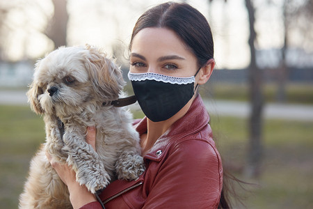 在检疫期间，人们在户外用面罩拍下了一张漂亮而细心的黑发横向照片，带着心爱的宠物散步，牵着狗，直接看着相机。待在家里的概念.