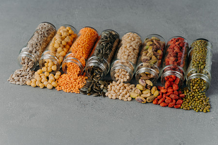 Dried seeds, beans and various grains poured out from glass bottles, spread on grey background. Healthy ingredients for your nutrition. Organic food