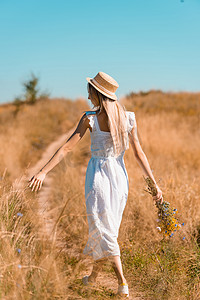 在草地上，穿着白衣、头戴草帽、手持野花走在路上的年轻女子的背影