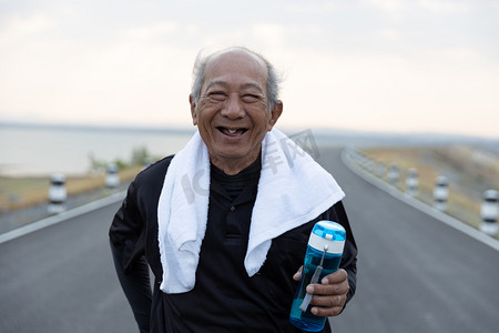 前瞻亚洲老年人运动快乐的微笑,积极向上的情感.在早晨的阳光下,把老人抱在路上的水瓶关上.健康生活方式和运动概念.