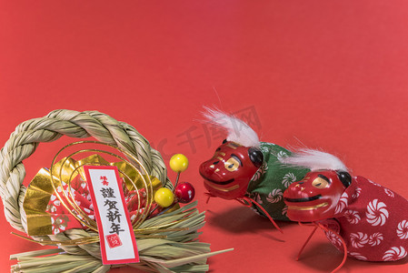 cone摄影照片_Japanese New Year's Cards with black ideograms Gingashinnen which means Happy New Year with cute Japanese Folklore  animals figurines of two lions Shishimai, origami made range and straw ornament with pine cone on red background. It's also write in r