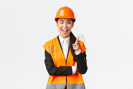 指指点点摄影照片_穿着安全帽和反光夹克，微笑专业的亚洲女施工经理，招揽新大楼的入住、眨眼和对着相机指指点点，向客户展示最佳地产
