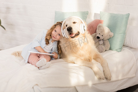 躺着的人像摄影照片_可爱的孩子拿着平板电脑, 靠在金毛猎狗身上, 耳机躺在儿童房的床上