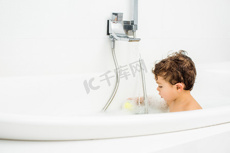 洗澡玩具摄影照片_幼儿男孩坐在白色浴室, 玩洗澡玩具