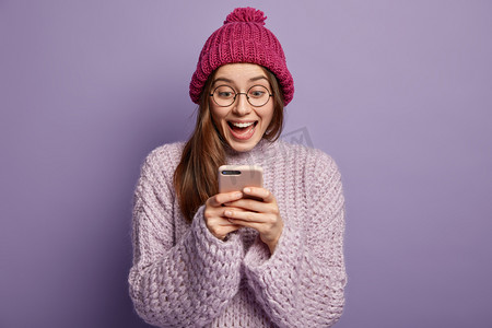 uses摄影照片_Happy female blogger uses modern gadget for browsing internet, mobile phone application, gazes with smile at screen, reads good news online, wears pink headgear with pompon, warm oversized jumper