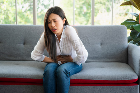 肚子痛摄影照片_不快乐的亚洲女人坐在沙发上，忍受着肚子痛。月经、腹泻或消化不良引起的腹痛。疾病和保健概念