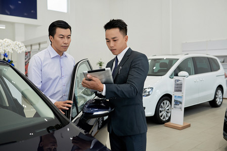 员工电脑摄影照片_在咨询客户时，年轻的亚洲汽车经销店员工在平板电脑上阅读有关汽车的信息