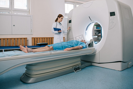 年轻放射技师在给病人做断层扫描准备的同时操作CT扫描仪