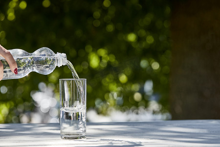 在阳光明媚的日子里，女人在木制桌子上用透明的杯子从瓶子里倒出新鲜的清洁水的剪影